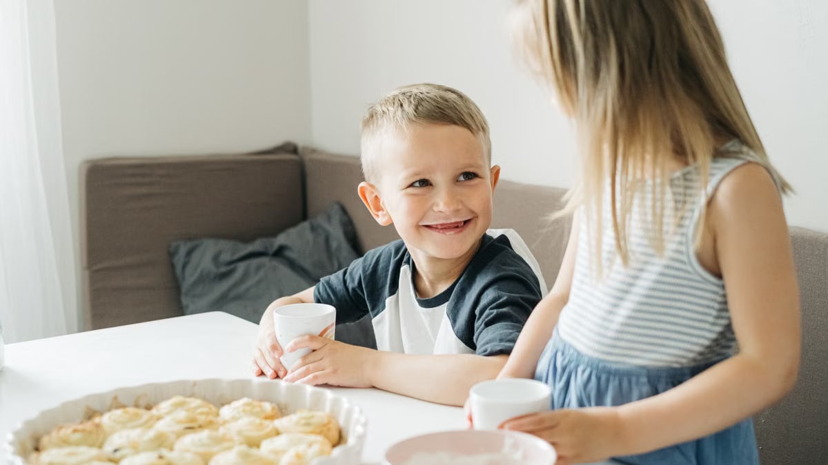 Smiling siblings sharing a breakfast table with milk and pastries in a cozy home setting.